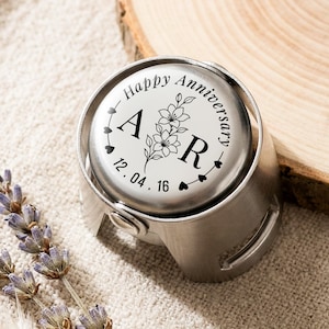 May include: A circular silver-toned metal bottle stopper featuring a white top insert engraved with the text Happy Anniversary, the letters A and R, a floral illustration, and the date 12.04.16, accented by a series of small heart symbols.
