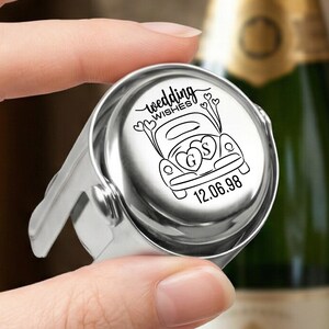 May include: A silver-toned metal bottle stopper featuring a circular top engraved with a black line drawing of a car, heart-shaped balloons, the text "wedding WISHES", the initials "G" and "S" inside hearts, and the date "12.06.98".