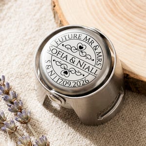 May include: A silver-toned metal bottle stopper featuring a circular top engraved with the text FUTURE MR & MRS, SOFIA & NIALL, and the date 17.09.2026, accented with decorative scrollwork and heart motifs centred on the flat circular surface.