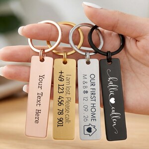 May include: Four rectangular metal keychains in rose gold, gold, silver, and black finishes. Each features a split ring and custom engraving: "Your Text Here," "I am lost. Please call: +49 123 456 78 901," "OUR FIRST HOME M & B 12.09.26," and "hallie cullen" with a heart icon.