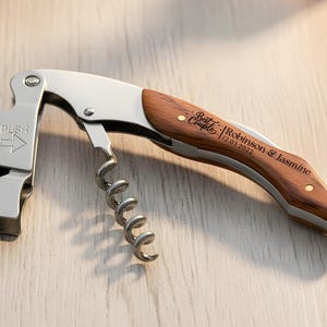 May include: A stainless steel waiter's corkscrew with a wooden handle inlay. The handle is engraved with the text "Best Couple" followed by "Robinson & Jasmine" and the date "12.03.2022". The metal lever features the word "PUSH" and an arrow.