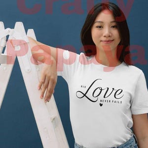 May include: A white t-shirt with the text "His Love Never Fails" in black script. The shirt is paired with blue denim jeans. The image features a woman leaning against a white ladder.