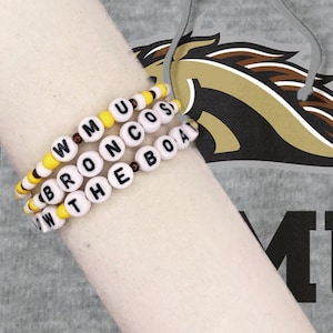 May include: Three beaded bracelets spelling out "WMU BRONCOS ROW THE BOAT" in white letter beads. The bracelets have yellow and brown accent beads. The background includes a gray shirt with a Bronco logo.