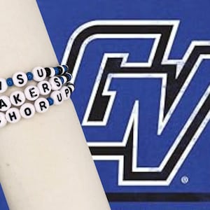 May include: A white beaded bracelet with the words "GVSU Lakers Anchor Up" in black letters. The bracelet is on a person's arm in front of a blue background with a white and black "GV" logo.