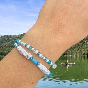 May include: Two beaded bracelets on a wrist. The top bracelet is white and blue with iridescent beads. The bottom bracelet is blue with white letter beads that spell "LAKE LIFE".
