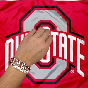 Könnte beinhalten: Zwei Armbänder mit Perlen, die "OSU OHIO BUCKEYE" mit weißen Buchstabenperlen buchstabieren, mit roten und schwarzen Akzentperlen. Die Armbänder befinden sich an einem Handgelenk, vor einem roten Hintergrund mit einem großen weißen und grauen Logo.