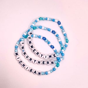 Peut inclure: Trois bracelets en perles avec les mots "SUMMER TIME", "NORTHERN" et "MICHIGAN" écrits en lettres noires sur des perles blanches. Les bracelets sont faits de perles blanches et bleues, dont certaines sont bleu clair et turquoise.