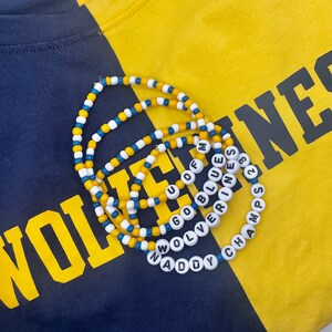 May include: Three beaded necklaces with the words "Go Blue", "Wolverine", and "Naddy Champs 23" on a yellow and blue shirt. The shirt has the word "WOLVERINES" printed on it.