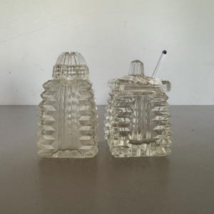 May include: A clear glass salt and sugar set. The salt shaker is a tall, faceted bottle with a domed lid. The sugar bowl is square with a lid and a small spoon. The set is displayed on a neutral surface.