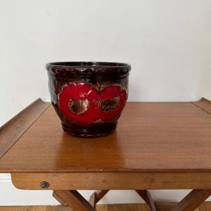 May include: A brown ceramic plant pot with a glossy finish. The pot has a striking red floral design on the front. The cylindrical pot sits on a wooden table. A decorative piece for indoor plants.