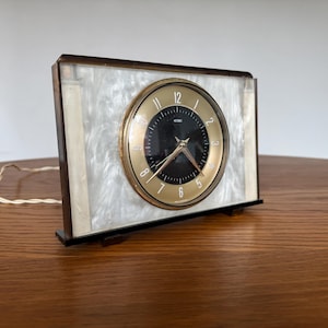 May include: A retro electric clock with a gold-coloured face and black numerals. The clock is set in a rectangular frame with a pearlescent white and brown border. The clock has a black base and a power cord.