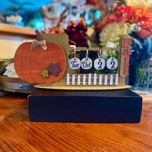 May include: Wooden fall-themed calendar with a pumpkin, a white picket fence, and a sign that says "Fall". The calendar has a date display that says "Tue Oct 2 2022".