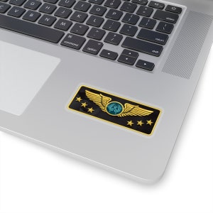May include: A gold and black video game rank badge with five stars and a pair of wings. The wings have a green and blue emblem in the center.