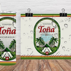 May include: A tumbler and a poster featuring the "Cerveza Toña Nicaragua" design. The design includes the brand name in red, palm trees, and a road leading to a mountain. The tumbler has a straw and the poster is held by a clip.