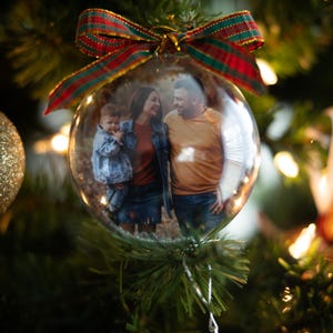 May include: A clear glass Christmas ornament with a family photo inside, hanging on a decorated tree. The ornament is topped with a red, green, and gold ribbon bow. The tree has lights and other ornaments.