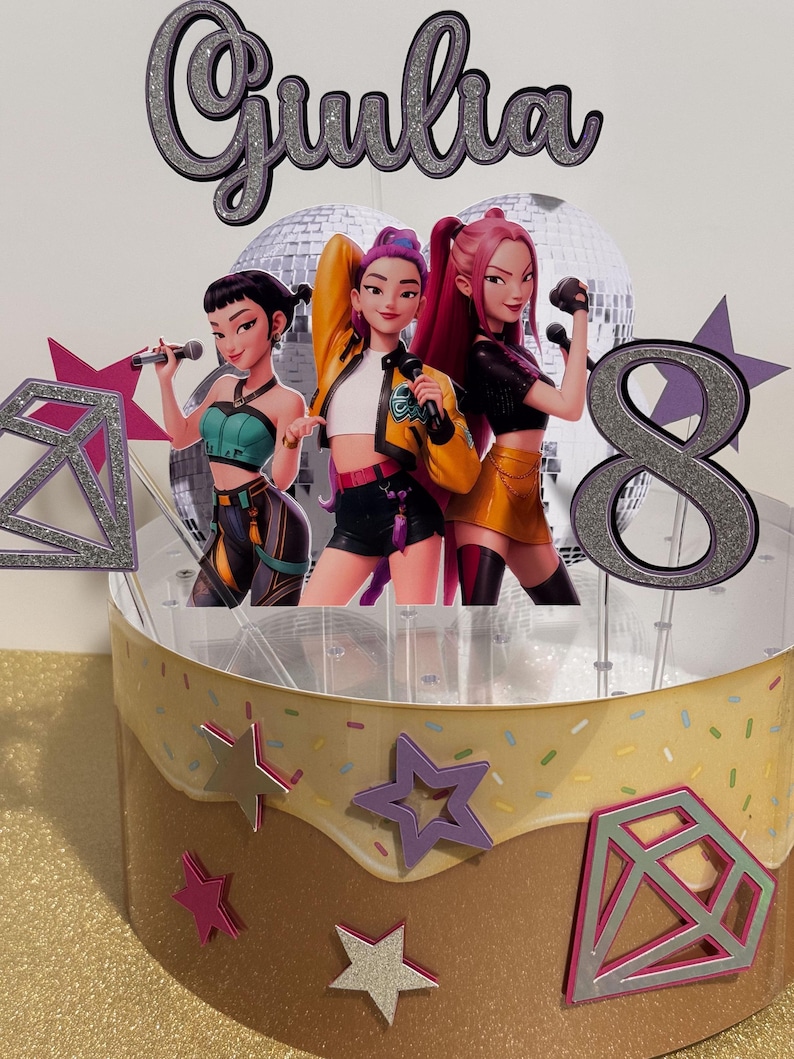 May include: A birthday cake topper featuring three animated female singers with microphones. The topper includes the name "Giulia" and the number "8" in silver glitter. The cake is decorated with star and diamond shapes.