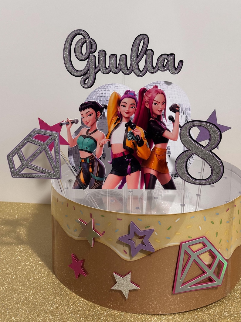 May include: A custom cake topper featuring three animated female figures with microphones, the name "Giulia", and the number "8". The cake is decorated with star and diamond shapes, and has a brown and tan frosting design.