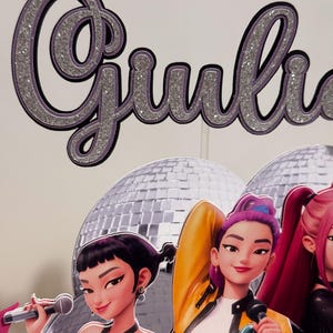 May include: A glittery silver and purple "Giulia" cake topper with a black outline. Below, three animated female singers with microphones stand in front of a disco ball backdrop. The singers wear colorful outfits.