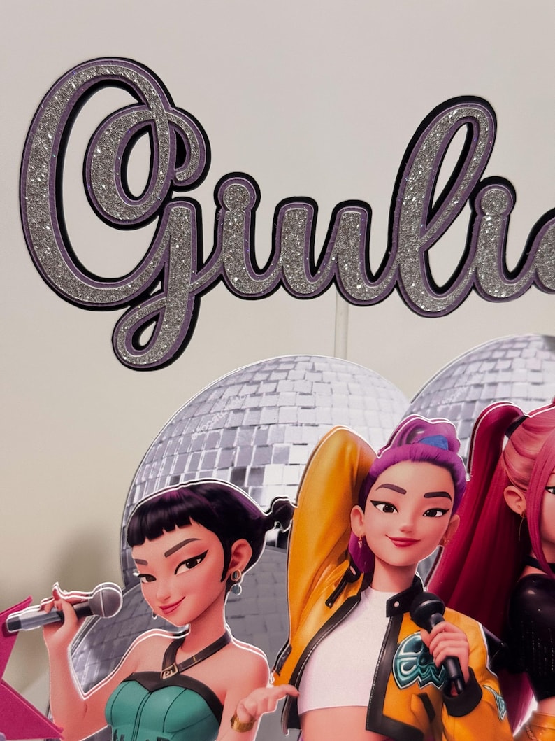 May include: A glittery silver and purple "Giulia" cake topper with a black outline. Below, three animated female singers with microphones stand in front of a disco ball backdrop. The singers wear colorful outfits.