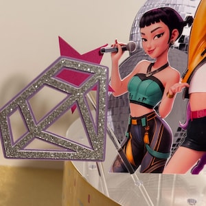 May include: A cake topper featuring a cartoon woman holding a microphone in front of a disco ball. The woman is wearing a teal top and black pants. A purple diamond-shaped decoration with silver glitter accents is in the foreground.