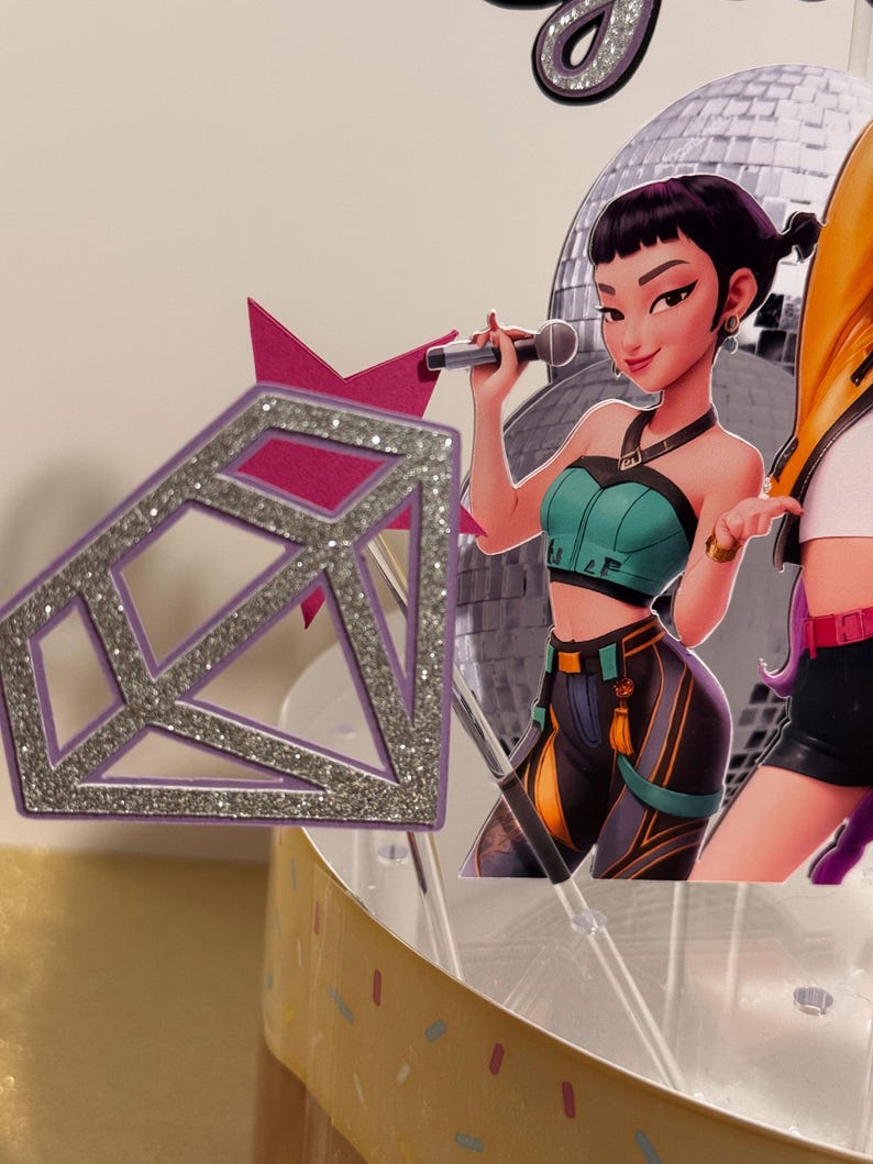 May include: A cake topper featuring a cartoon woman holding a microphone in front of a disco ball. The woman is wearing a teal top and black pants. A purple diamond-shaped decoration with silver glitter accents is in the foreground.