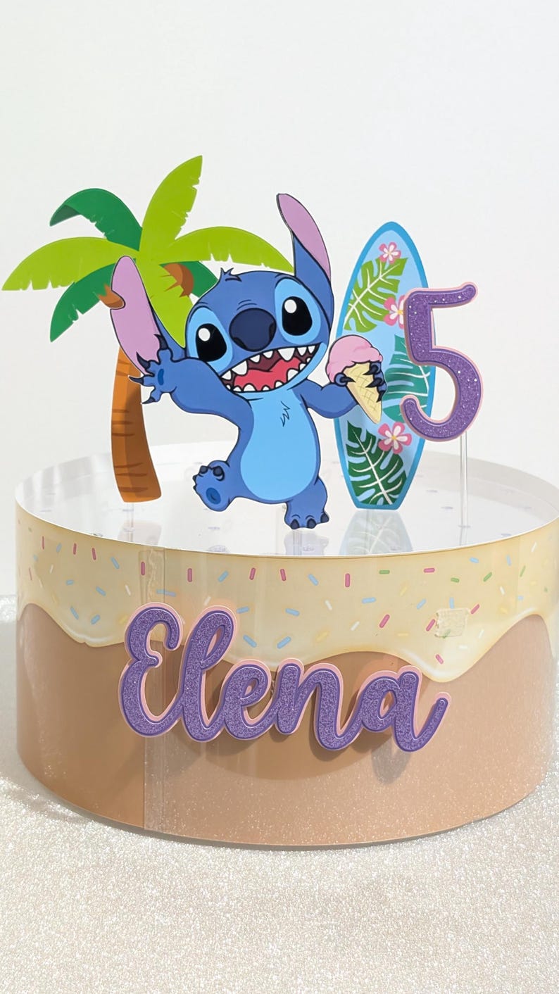 May include: A cake with a cartoon character, palm tree, surfboard, and the number 5. The cake has a light brown and beige frosting with sprinkles and the name "Elena" in purple script.