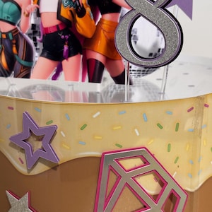 May include: A cake topper featuring a silver glitter number 8, with a purple outline. The cake is decorated with a star and diamond shape, and images of animated characters. The cake has a brown and yellow frosting.