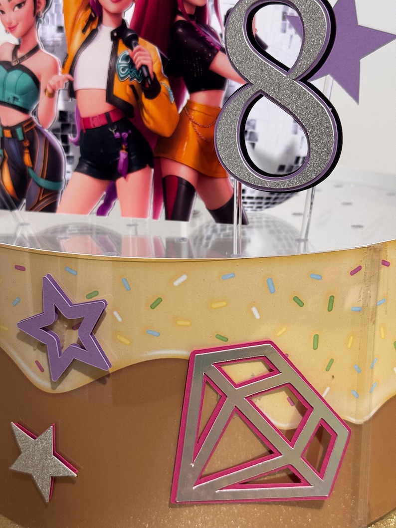 May include: A cake topper featuring a silver glitter number 8, with a purple outline. The cake is decorated with a star and diamond shape, and images of animated characters. The cake has a brown and yellow frosting.