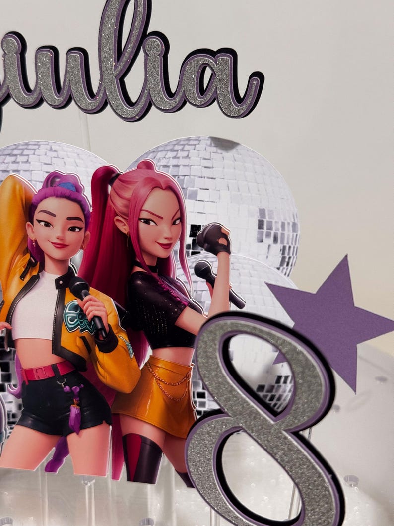 May include: A cake topper featuring two animated female singers with microphones, a silver glitter number 8, and the name "Julia" in silver glitter. The singers are in colorful outfits, with disco balls in the background.