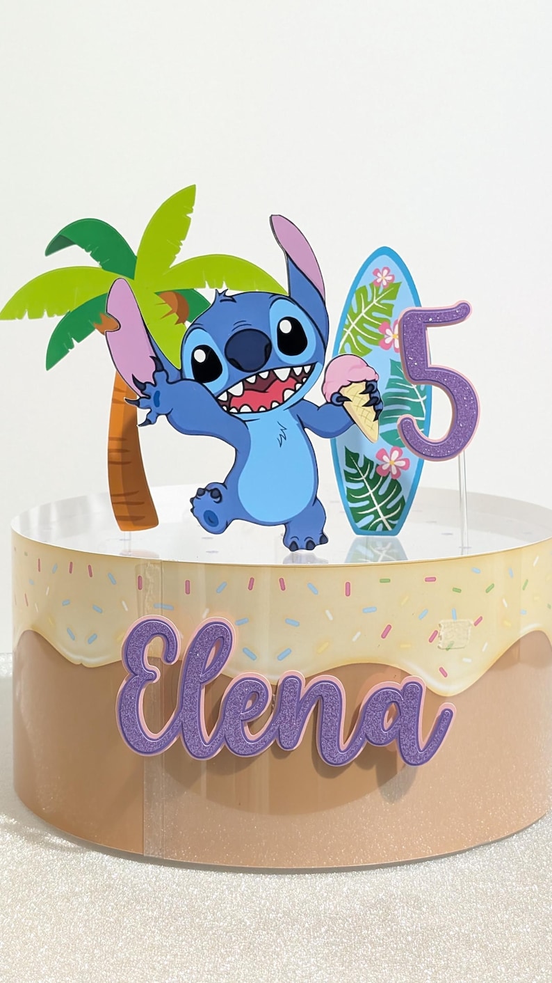 May include: A cake topper featuring the character Stitch, a surfboard, a palm tree, and the number 5. The cake is decorated with the name "Elena" and sprinkles. The cake is designed for a birthday celebration.