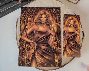 May include: A gold bookmark with a woman in a gold dress standing in front of a gold background. The bookmark has the text "BRILLO DESTELLO LA PRISIONERA DE ORO" on it.