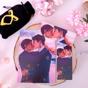 May include: Two men in suits are kissing on a beach with a pink sky. The image is printed on a white card with a gold border. The card is on a wooden surface.