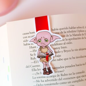 May include: A red bookmark featuring a cartoon illustration of a pink elf holding a book. The elf has green eyes and a worried expression. The text on the bookmark reads: "Hunt - Ho - Se fu - Ruh. - El I del Cuerno. Ella lev - ¿Se h La sonrisa amarga de Ruhn no la consoló. -Me ha advertido del <<veneno>>> que escu -Debo tomarmelo como un cumplido, s Ruhn no sonrió en esta ocasión. -Quiere saber lo que harás con el Cuer -Lo usaré como mi nueva jarra para los Hustcoltó una risotada al entrar de nue Lament"