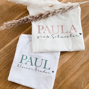 Könnte beinhalten: Zwei weiße T-Shirts mit aufgedrucktem Text. Das obere Shirt sagt "PAULA große Schwester" mit einem rosa Herzen. Das untere Shirt sagt "PAUL kleiner Bruder" mit einem grünen Stern.