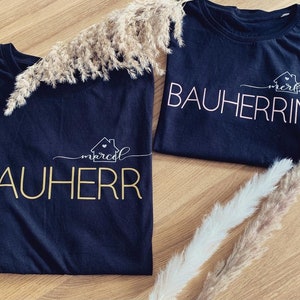 Könnte beinhalten: Zwei schwarze T-Shirts mit dem Text "Bauherr" und "Bauherrin" in goldener und weißer Schrift. Die Shirts liegen auf einer Holzoberfläche mit getrocknetem Gras.