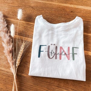 Könnte beinhalten: Weißes T-Shirt mit dem Wort "FÜNF" in bunten Buchstaben geschrieben, darunter das Wort "frieda" in Kursivschrift.