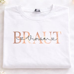 Könnte beinhalten: Ein weißes T-Shirt mit dem Wort BRAUT in einer metallisch roségoldenen Schriftart. Der Name Vivian ist in schwarzer Schreibschrift über die Mitte des Wortes geschrieben, wobei kleine Herzformen in die Buchstaben integriert sind.