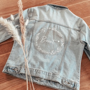 Könnte beinhalten: Eine hellblaue Jeansjacke mit einem weißen Blumengirlanden-Design auf der Rückseite. Die Girlande hat das Wort "Braut" darin geschrieben.