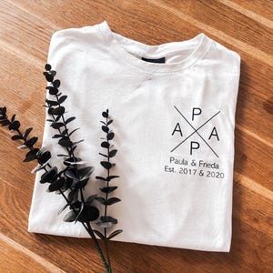 Könnte beinhalten: Weißes T-Shirt mit einem schwarzen Grafikdesign mit den Buchstaben P, A und einem zweiten P und A, angeordnet in Kreuzform. Darunter steht der Text "Paula & Frieda Est. 2017 & 2020". Das Shirt liegt gefaltet auf einer Holzoberfläche, mit schwarzen Zierblättern.