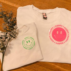 Könnte beinhalten: Zwei weiße T-Shirts mit einem Smiley-Motiv. Ein Shirt hat ein pinkfarbenes Smiley mit dem Text "HAPPY HAPPY HAPPY" in Pink. Das andere Shirt hat ein grünes Smiley mit dem Text "HAPPY HAPPY HAPPY" in Grün.
