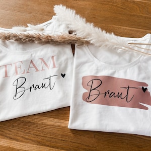 Könnte beinhalten: Zwei weiße T-Shirts mit dem Aufdruck "Team Braut" und "Braut". Der Text ist in Schwarz und Rosa mit einem kleinen schwarzen Herz neben jedem Wort.