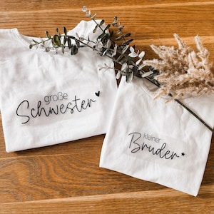 Könnte beinhalten: Zwei weiße T-Shirts mit schwarzem Text, der "große Schwester" und "kleiner Bruder" lautet. Die Shirts sind mit einem kleinen Herz und Stern verziert. Die Shirts liegen auf einer Holzoberfläche.