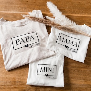 Könnte beinhalten: Drei weiße T-Shirts mit schwarzem Text: "PAPA", "MAMA" und "MINI" in rechteckigen Kästen, mit einem Herz und "EST. 2023" darunter. Die Shirts liegen auf einer Holzoberfläche, mit dekorativen getrockneten Pflanzen.