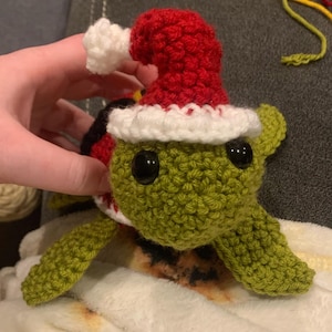 May include: A crocheted turtle figurine with a festive Christmas hat. The turtle is primarily green with black eyes and a red and white hat. The hat has a white trim and a white pom-pom. The turtle is resting on a white surface.