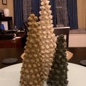 May include: Three handmade crochet Christmas trees in varying sizes and colors. The trees are in shades of brown, cream, and green. The trees have a textured, bobble stitch design, and are displayed on a white surface.