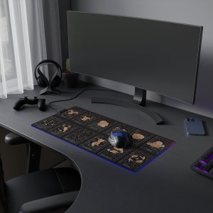 Puede incluir: Un escritorio de ordenador negro con un monitor de ordenador negro, unos auriculares de juego negros, una alfombrilla de ratón negra y dorada con un diseño zodiacal, un ratón de juego negro y azul, un teléfono inteligente negro y un teclado negro.
