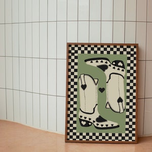 May include: A framed print with a checkered border and a green background. The print features two white cowboy boots with black hearts on them.