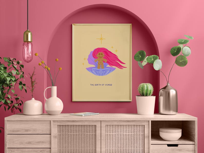 Op de afbeelding: Een digitale illustratie van de geboorte van Venus, een figuur met roze haar dat uit een schelp komt. De illustratie is ingelijst en hangt aan een roze muur boven een houten kast.
