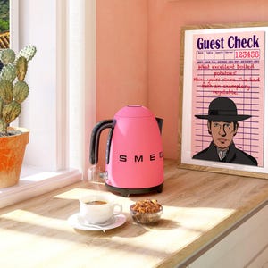Puede incluir: Una tetera eléctrica rosa con asa y pico negros, sentada sobre una encimera de madera junto a una taza de café, un pequeño cuenco de aperitivos y una impresión enmarcada con el texto "Guest Check 123456 What excellent boiled potatoes! Many years since I've had such an exemplary vegetable."