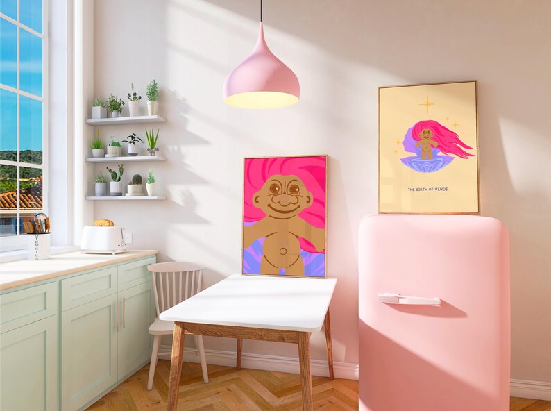 Op de afbeelding: Een roze keuken met een witte tafel en stoelen, een roze koelkast en twee ingelijste prints van een cartoon-troll met roze haar. De tekst op een print luidt "The Birth of a Friend".